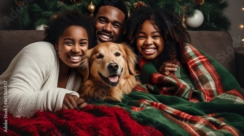 An African-American family with a golden retriever dog sit, covered with a blanket, on the sofa in the living room on Christmas Day