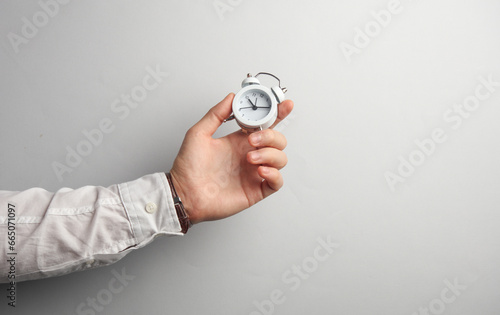 Fototapeta Naklejka Na Ścianę i Meble -  Man's hand in white shirt holds alarm clock on gray background