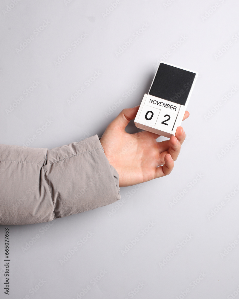 Man's hand in warm down jacket holds block calendar with date november 2 on gray background Man's hand in warm down jacket holds block calendar with date november 2 on gray background