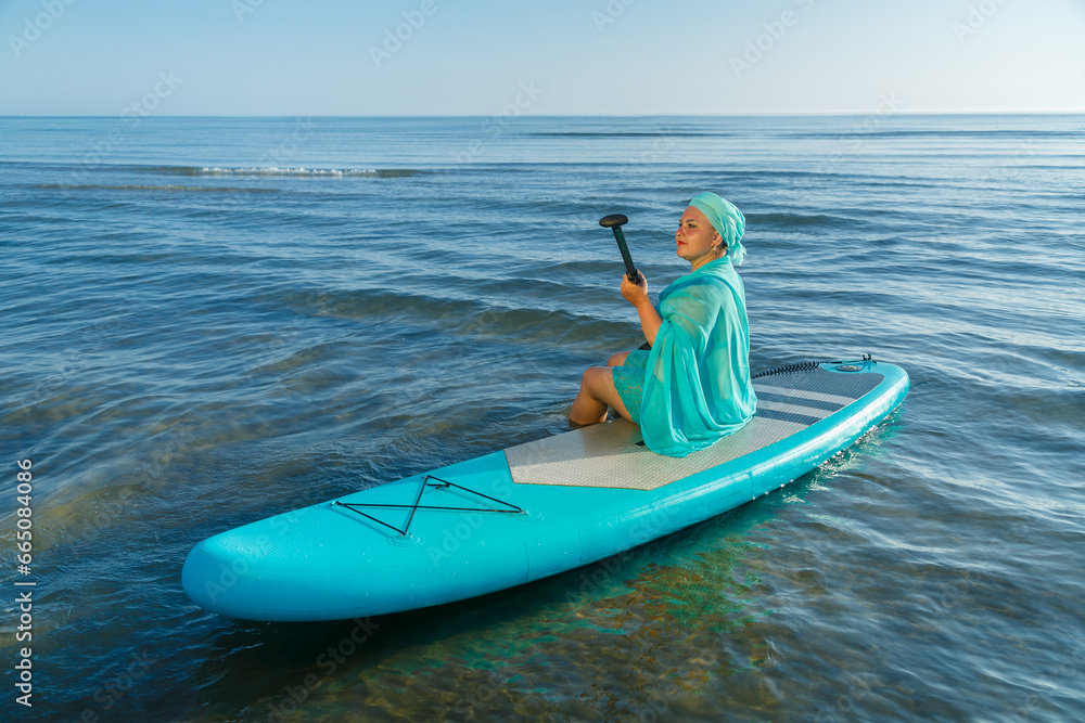 A Jewish woman in a turquoise swimsuit with a skirt and a scarf on her head kneeling on a SUP board near the sea.