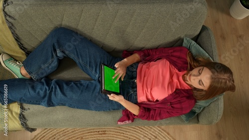 Top view capturing a young woman laying on the couch, looking at the tablet screen with an advertising area, workspace mock up on it as if she is searching for something online. photo