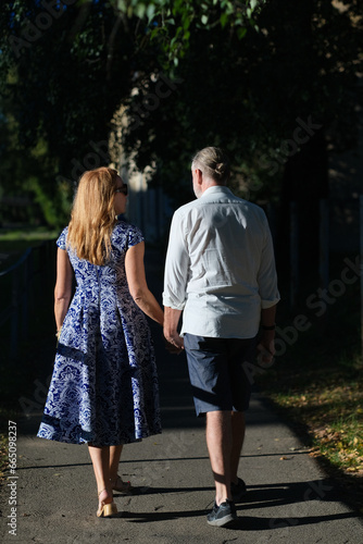 Woman and man full length from the back. The woman is wearing a blue dress with a white pattern and beige shoes. The man is wearing a white shirt, blue shorts and sports shoes. The city in the backgro