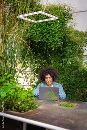 Smiling young ethnic guy ,dressed in casual clothes, works on a laptop outdoors, in a green area in nature. Green open space for coworking, freelancer, relaxing, coffee, food. Free place to work © SNAB
