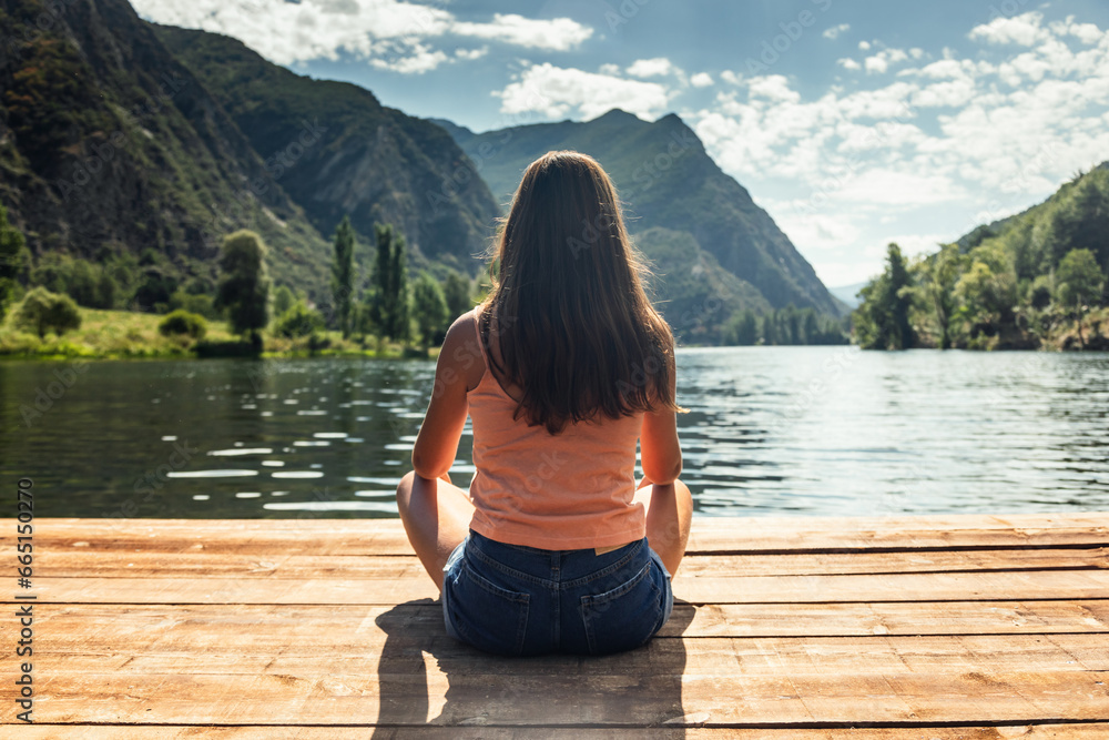 Back view of young woman relaxing while sitting near the lake in a mountain landscape Back view of young woman relaxing while sitting near the lake in a mountain landscape