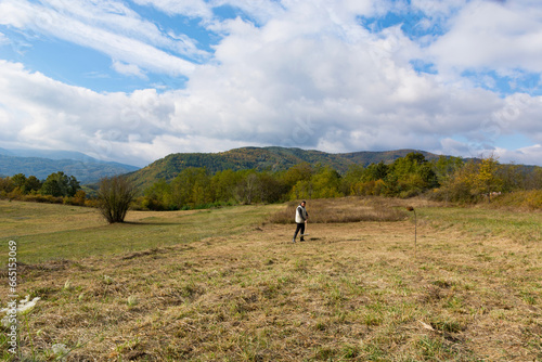 Autumn landscape with meadows and a farmer gathering hay with a rake. In the background, the forest and hills have taken on autumn colors. The day is variably cloudy © Emilija