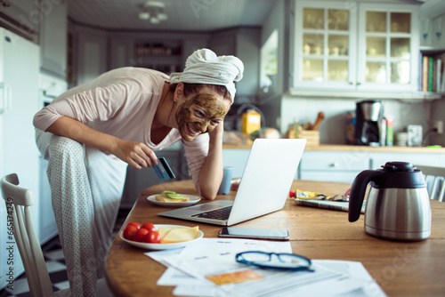 Attractive young woman with a facial mask using credit card on the laptop in he kitchen