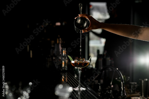 Selective focus of backlited martini glass in which female bartender pours liquid from steel shaker cup