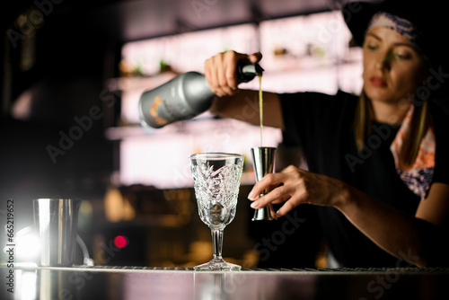 View of girl bartender at bar counter holding bottle vith syrup and pours it to steel jigger for making cocktail