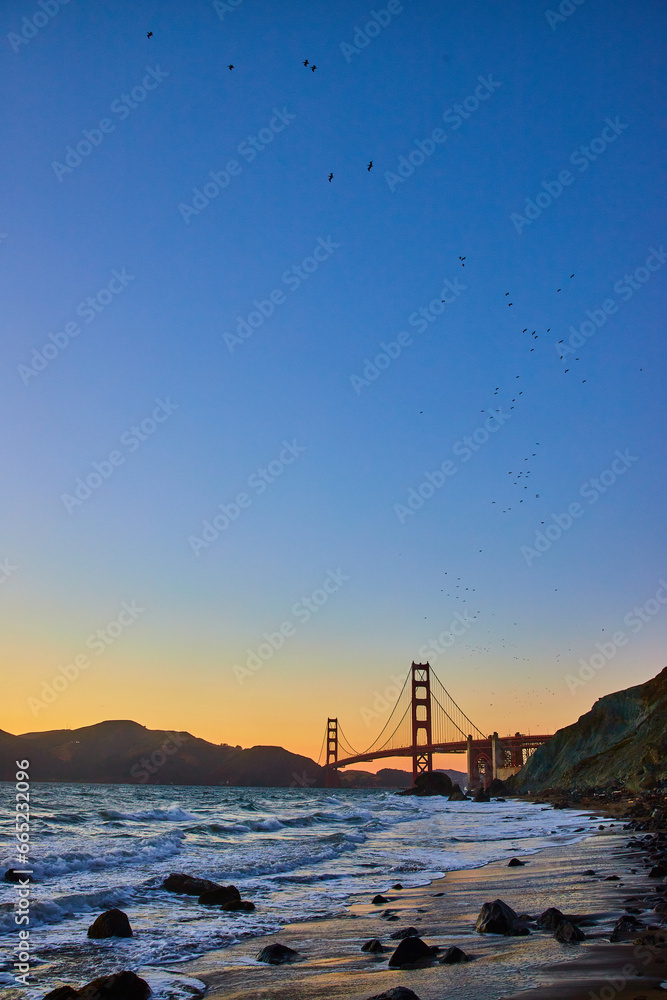 Sunset sky above glowing blue waves and Golden Gate Bridge along sandy shore