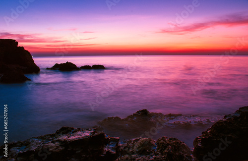 Along the coast there are rocks and sea waves with the sun setting behind the horizon. There are dark rocks because there is little light. and the gentle waves of the sea in summer