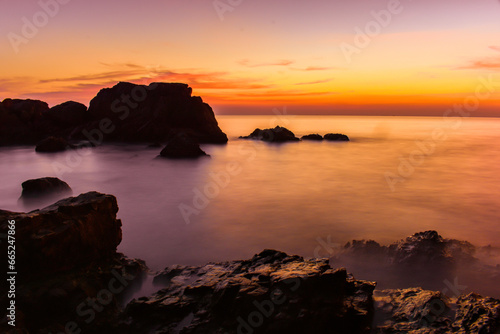 Along the coast there are rocks and sea waves with the sun setting behind the horizon. There are dark rocks because there is little light. and the gentle waves of the sea in summer