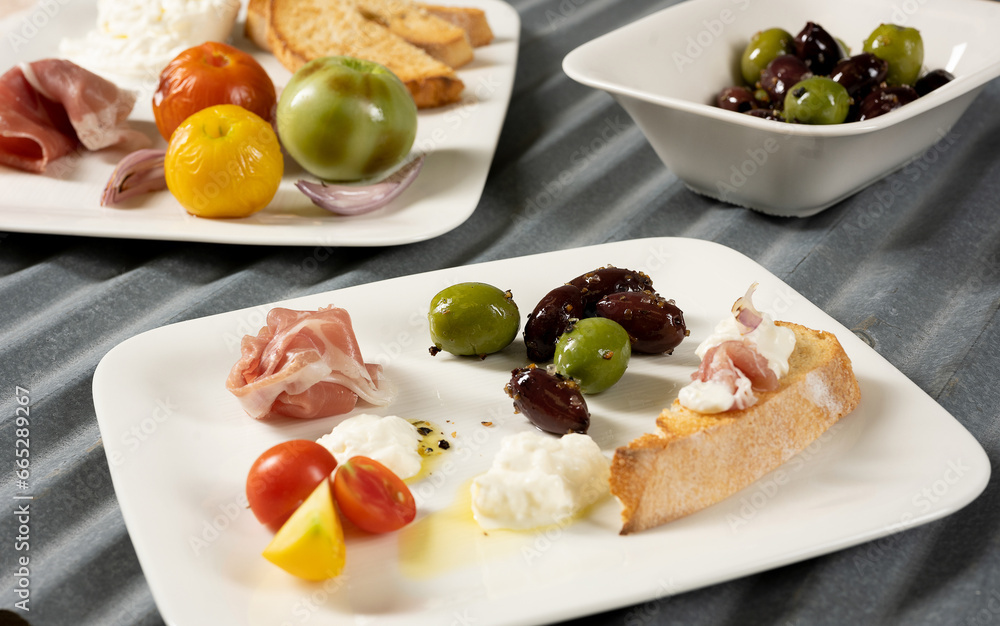 Multiple appetizers on white plates with roasted tomatoes, olives, prosciutto, olive oil, bread Multiple appetizers on white plates with roasted tomatoes, olives, prosciutto, olive oil, bread