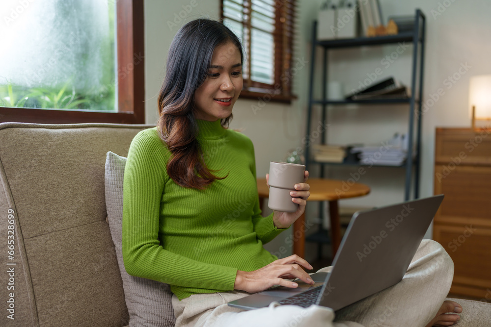 Young successful professional Asian businesswoman in casual clothes sits on a sofa, reads a book, writes notes in a notebook while working on a laptop computer at home office in the living room. Young successful professional Asian businesswoman in casual clothes sits on a sofa, reads a book, writes notes in a notebook while working on a laptop computer at home office in the living room.