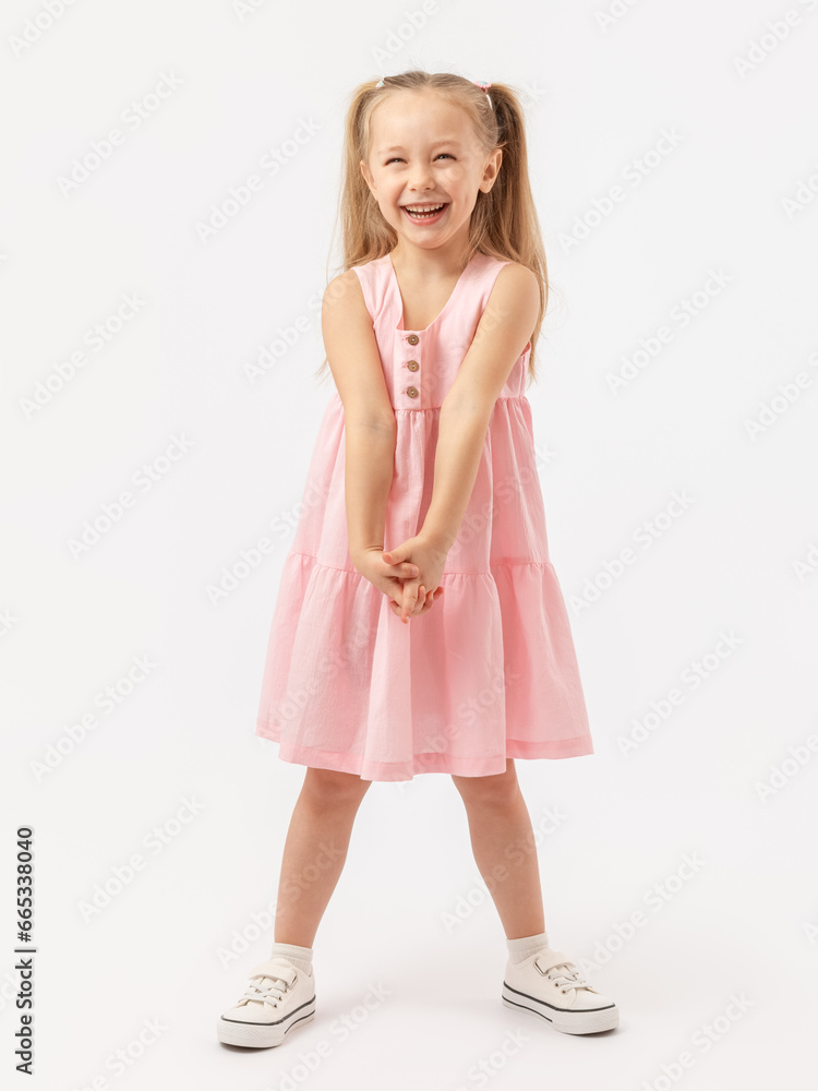A cheerful girl with long hair in a pink dress and sneakers stands on a white background and laughs sincerely.