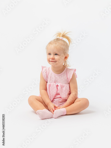 A cute blonde girl 1-2 years old in a pink dress is sitting on the floor smiling and looking away on a white background. Copy space.