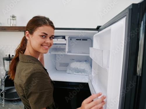 Woman smiling with teeth looking into camera in kitchen at home opened freezer empty with ice inside, home refrigerator, defrosted, view from back, stylish interior.