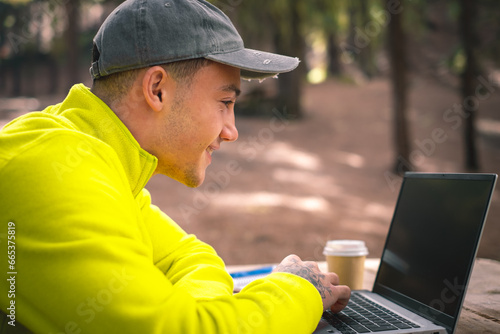Beautiful caucasian boy influencer working on laptop sitting outdoors in the forest. Hipster young man traveler working distantly while enjoying nature landscape during vacations photo