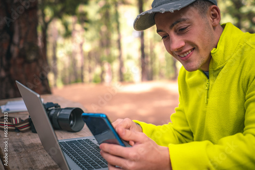 Young caucasian male freelancer using mobile phone while working on laptop computer remote. Hipster boy traveler working distantly while enjoying nature landscape during vacations