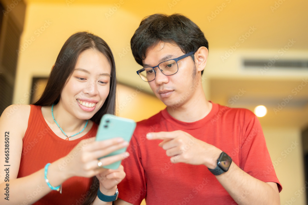 Close-up image of Asian businessman, man and woman, wearing red shirts. Smiling and pointing at smartphones together working on corporate online marketing work in a comfortable break room. Close-up image of Asian businessman, man and woman, wearing red shirts. Smiling and pointing at smartphones together working on corporate online marketing work in a comfortable break room.