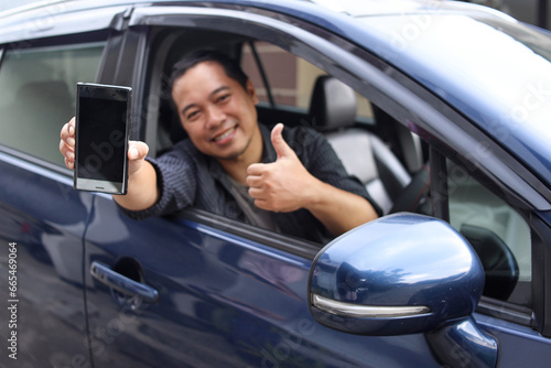 Male online taxi driver showing phone screen and thumb up while sitting in a car