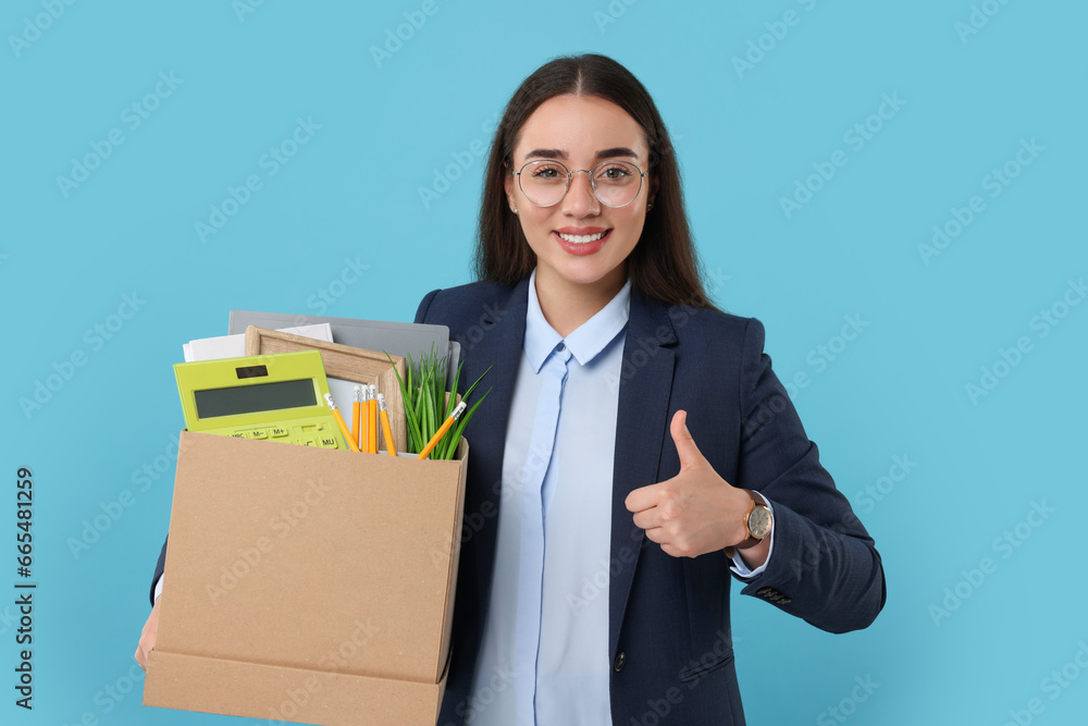 Happy unemployed woman with box of personal office belongings showing thumbs up on light blue background Happy unemployed woman with box of personal office belongings showing thumbs up on light blue background