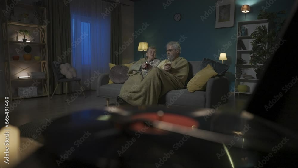 In the foreground frame, a vinyl turntable spins records. In the background an elderly couple sits under a plaid on a sofa with a glass of wine. HDR BT2020 HLG Material. In the foreground frame, a vinyl turntable spins records. In the background an elderly couple sits under a plaid on a sofa with a glass of wine. HDR BT2020 HLG Material.