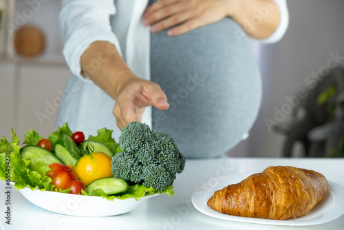 Pregnant woman choose croissant or fresh vegetables think about what to eat. The concept of healthy and unhealthy food, mindful eating during pregnancy. Diet while gestational diabetes. photo