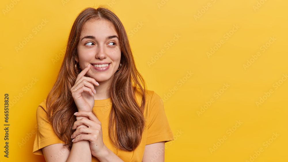 Horizontal shot of good looking young woman with long hair keeps hand near corner of lips concentrated aside smiles gently dressed in casual t shirt isolated over yellow background copy space for text - obrazy, fototapety, plakaty 
