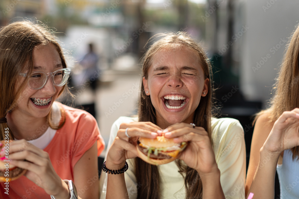 Portrait of joyful smiling teenage girls (14-15 years old) sitting at a cafe outdoors, enjoying hamburgers. Young ladies holding burgers, ready to eat them with pleasure.