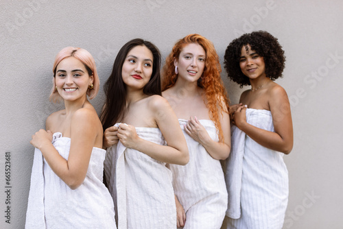 Four girls of different races, dressed in beige towels, stand against a grey wall. Beauty of diversity, SPA experience and multiethnic female friendship concept. Focus on first (Latin) girl.