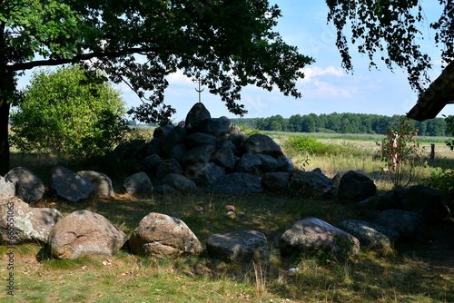 A close up on a stone circle made out of massive boulders, rocks, and stones with some Celtic cross on top of it located in a small forest or moor next to some fields and pasturelands in Poland