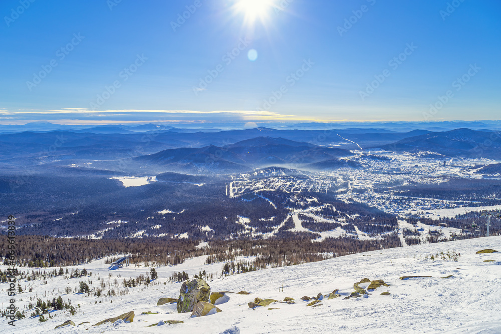 View from mount Mustag of slope in Sheregesh ski resort, Sheregesh village, picturesque panoramic landscape with range mountains and hills, forest, blue sky, sun and snow, sunny winter weather