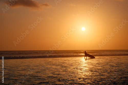 A Lone Surfer Carries His Board in the Shallow Waters, Bathed in Kuta's Cinematic Orange Sunset Glow
