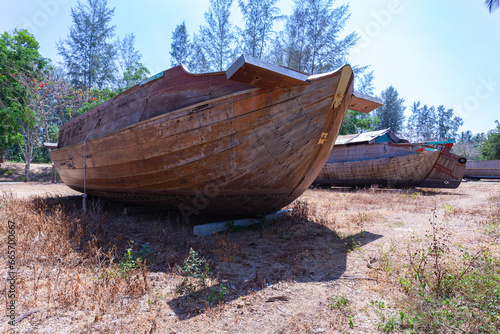 An ancient tugboat built of wood on the beach..A large wooden boat is displayed on the beach with a blue sky. .Line of pine trees along the beach background.