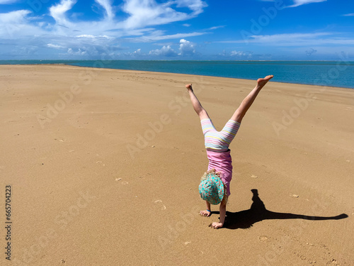 Young child wearing a blue sun hat and white shorts doing a cartwheel on an empty long sandy beach creating a dark shadow on the ground