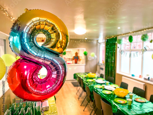 A room decorated for a child’s 8th birthday party with a large multicolored metallic foil, 8-shaped balloon and a long table with green table covering and party plates.