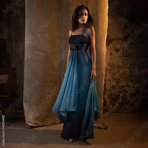 An elegant and beautiful brunette in a chic evening dress, a blue dress with a long flowing skirt to the floor. photo in dark colors on a textured wall, warm light
