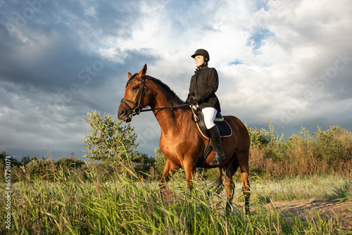 Horsewoman in equestrian sports gear, riding a horse, against an expressive sky, horseback riding in the open air © Ulia Koltyrina