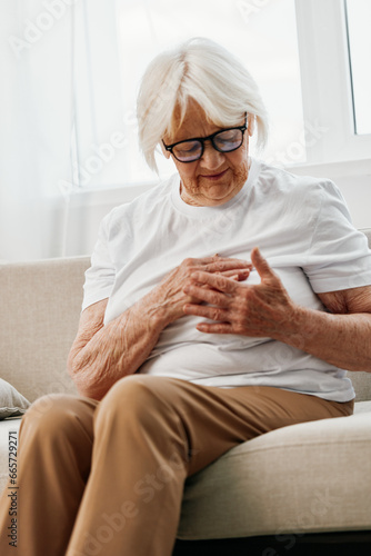 Elderly woman severe chest pain sitting on the sofa, health problems in old age, poor quality of life. Grandmother with gray hair holds her chest with her hands, women's health, breast cancer. © SHOTPRIME STUDIO