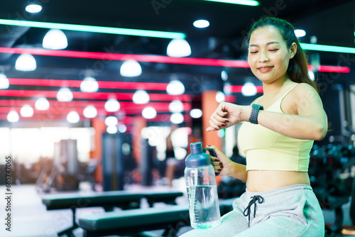 Good looking - beautiful Asian woman relaxing in the gym after made weight training and cardio workout, woman sitting or resting on the bench while drinking water after exercise.