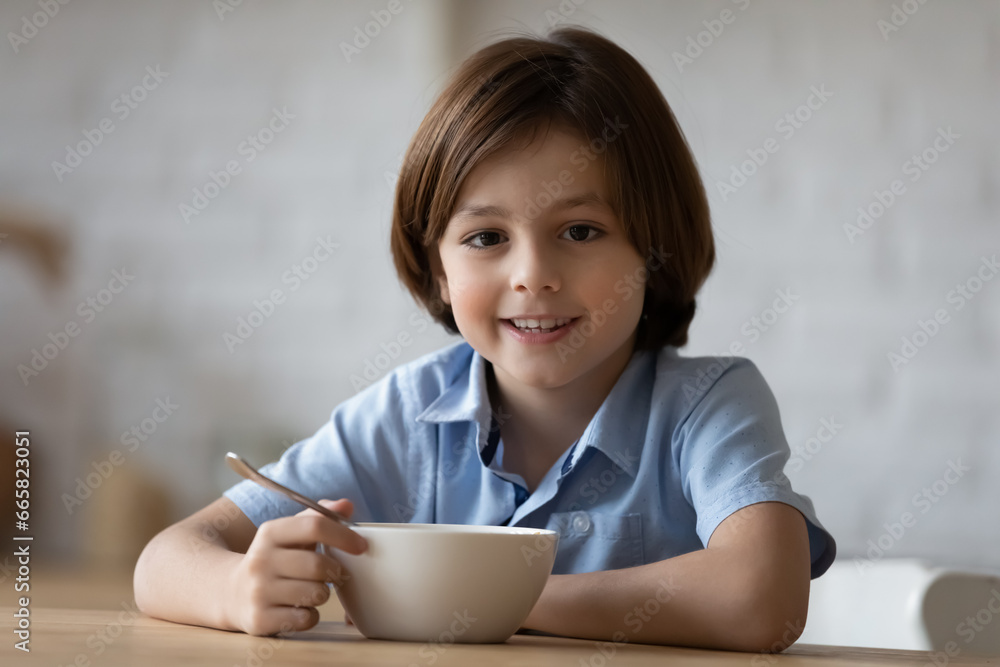Head shot portrait smiling 8s boy enjoying having breakfast in kitchen, happy adorable child kid looking at camera, sitting at table with tasty cereals or porridge bowl and spoon in morning Head shot portrait smiling 8s boy enjoying having breakfast in kitchen, happy adorable child kid looking at camera, sitting at table with tasty cereals or porridge bowl and spoon in morning