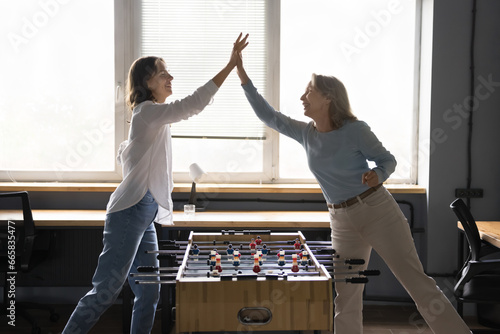 Young and mature women, two office employees playing table football, give high five, celebrate victory feel happy, enjoy lunch break together at workplace. Friendship, fun at workplace, companionship photo