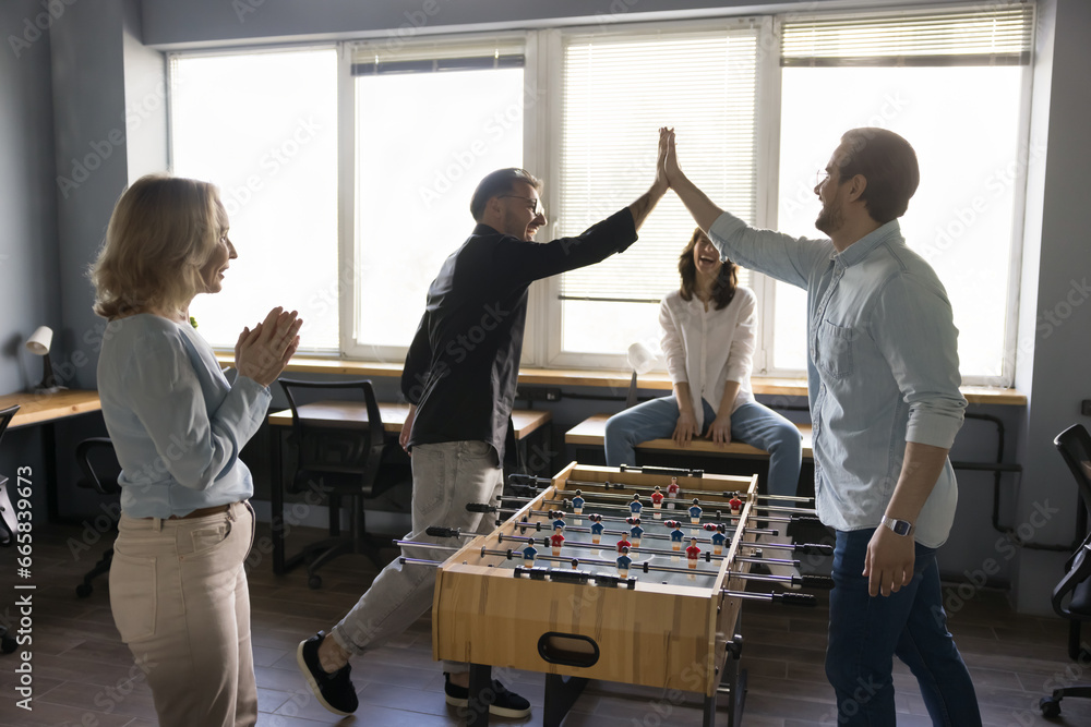 Group of businesspeople rest at lunch break play table foosball, have fun at workplace, enjoy teambuilding activity. Two young man colleagues give high five over board game celebrate final match over - obrazy, fototapety, plakaty 