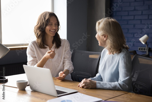 Two laughing colleagues, young and mature businesswomen talk, meet in office to solve business enjoy pleasant conversation distracted from workflow joking, looking cheerful and satisfied with teamwork