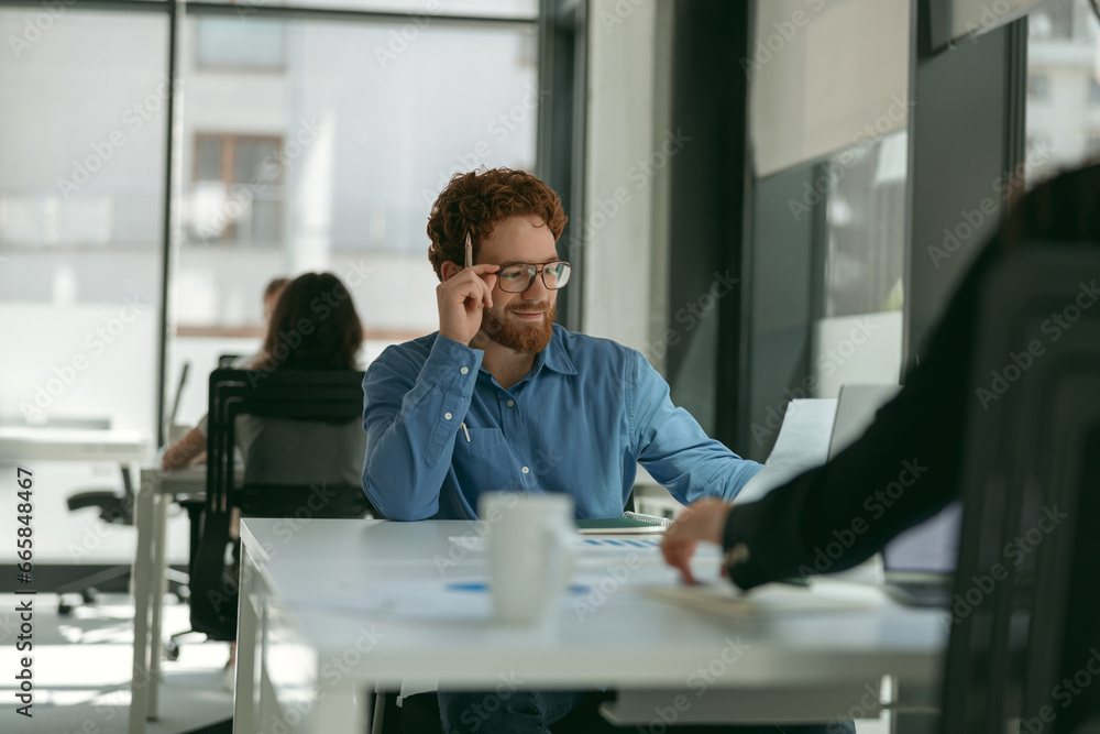 Smiling male sales manager in eyeglasses working on laptop sitting in office during working day Smiling male sales manager in eyeglasses working on laptop sitting in office during working day