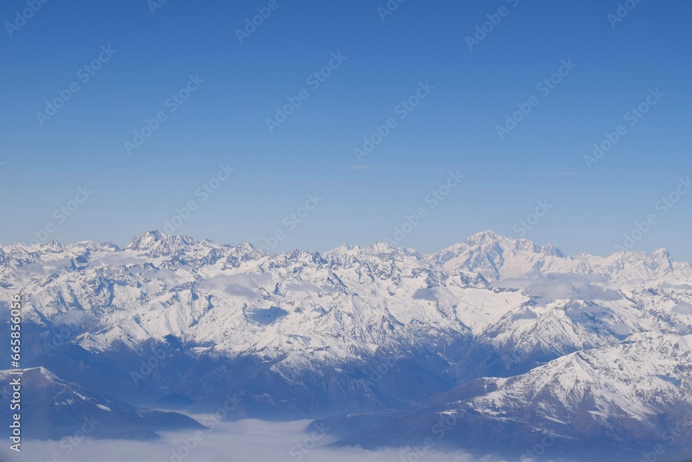 Beautiful aerial view of alpine snowcapped mountain range peaking through heavy clouds. Mountain peaks of Italian alps from above. The impressive winter view is taken from an airplane window. Beautiful aerial view of alpine snowcapped mountain range peaking through heavy clouds. Mountain peaks of Italian alps from above. The impressive winter view is taken from an airplane window.