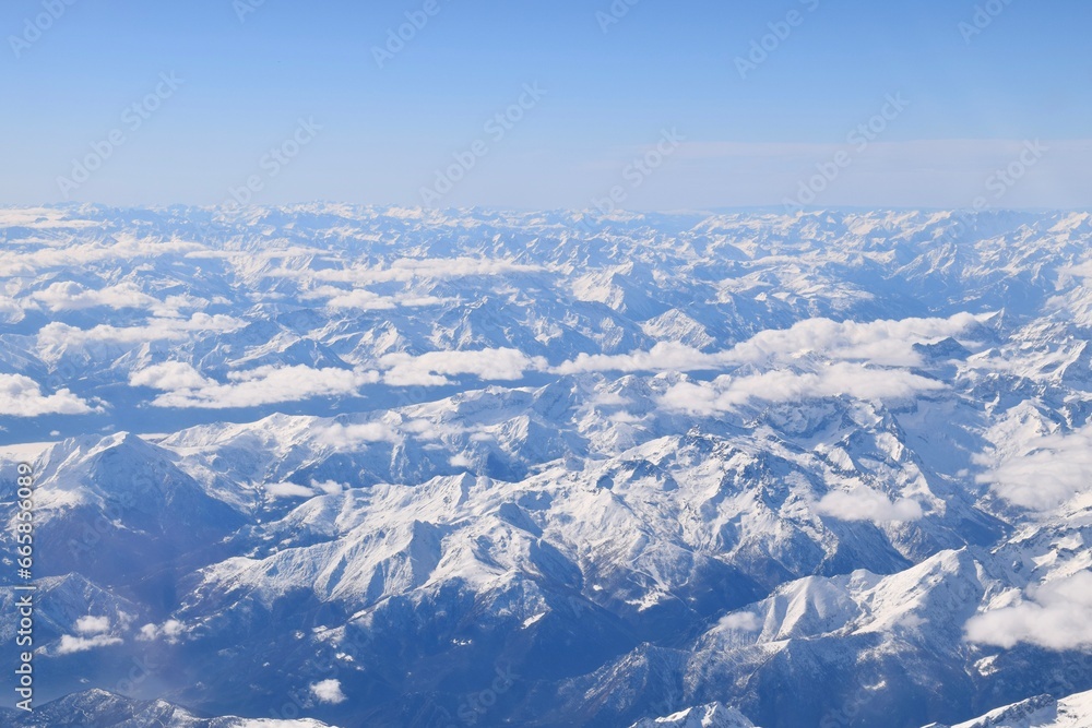Beautiful aerial view of alpine snowcapped mountain range peaking through heavy clouds. Mountain peaks of Italian alps from above. The impressive winter view is taken from an airplane window.