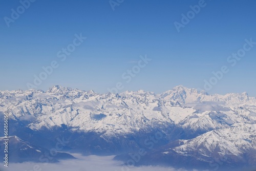 Beautiful aerial view of alpine snowcapped mountain range peaking through heavy clouds. Mountain peaks of Italian alps from above. The impressive winter view is taken from an airplane window.