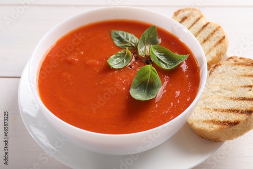 Delicious tomato cream soup in bowl with pieces of grilled bread on white wooden table, closeup