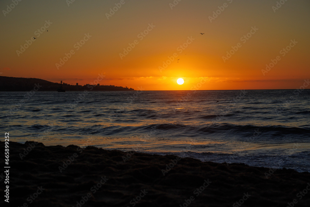 Atardecer en la costa de la ciudad, con el mar y sus olas, el sol se ve naranja en el horizonte